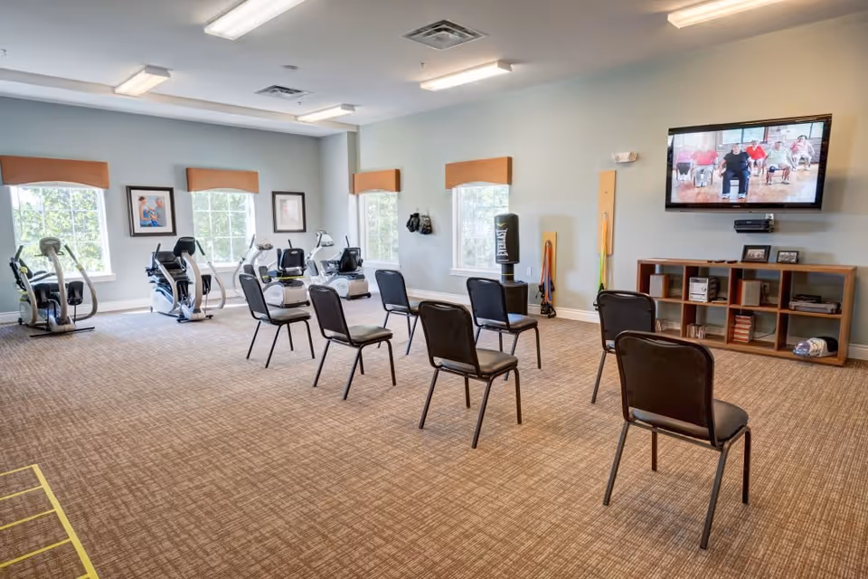 A spacious exercise room with several black chairs arranged in rows facing a wall-mounted TV screen showing a group exercise session. The room has large windows with orange valances, exercise bikes near the windows, a punching bag, and various exercise equipment along the walls. The floor is carpeted and the walls are painted light blue.