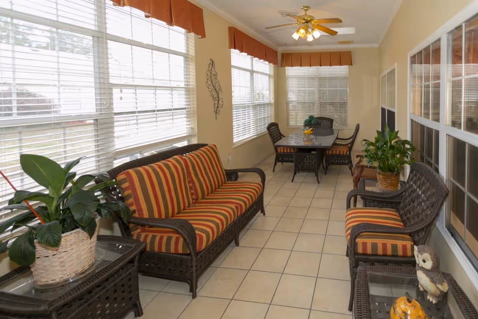 A bright sunroom with large windows covered by white blinds and orange valances. The room features wicker furniture with orange and brown striped cushions, including a sofa, two chairs, and a glass-top dining table with four chairs. There are potted plants on side tables and a ceiling fan with lights overhead. The floor is tiled in a light beige color.