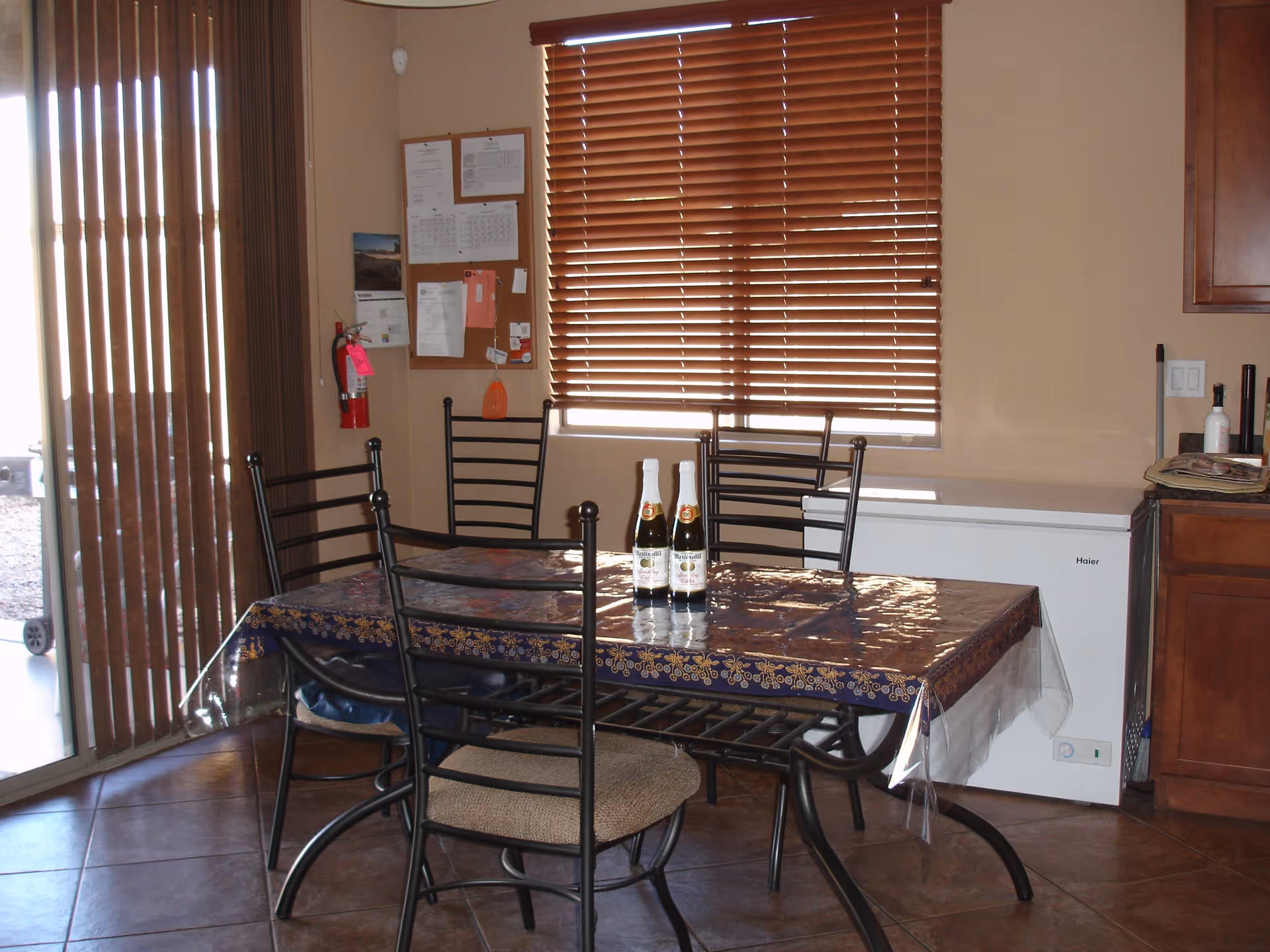 Dining area with a rectangular table covered with a plastic tablecloth and four metal chairs with cushioned seats. Two bottles are placed on the table. Behind the table is a window with closed wooden blinds, a bulletin board with papers pinned on the wall, and a fire extinguisher. To the right, there is a white freezer and wooden cabinets. Vertical blinds cover a sliding glass door on the left side.