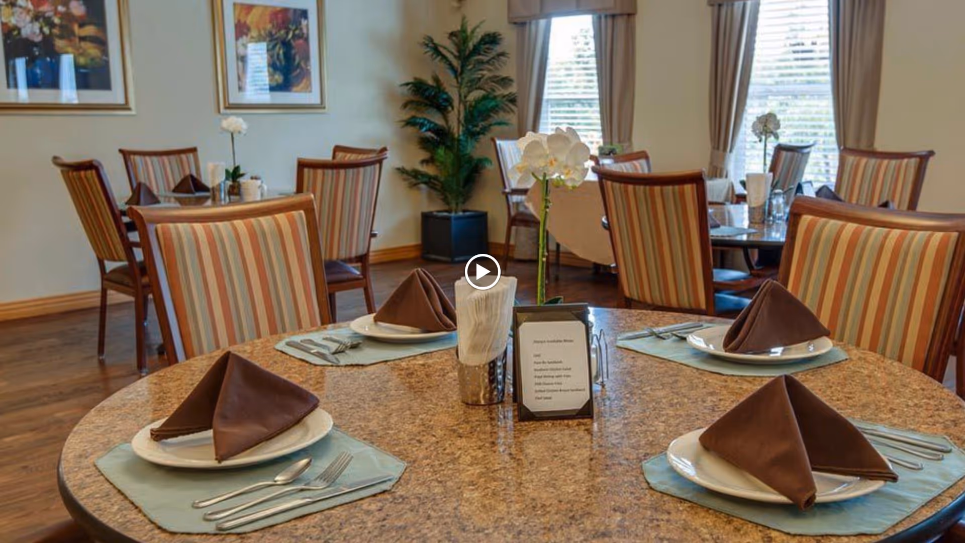 Dining room with round tables set with plates, folded brown napkins, and striped upholstered chairs.