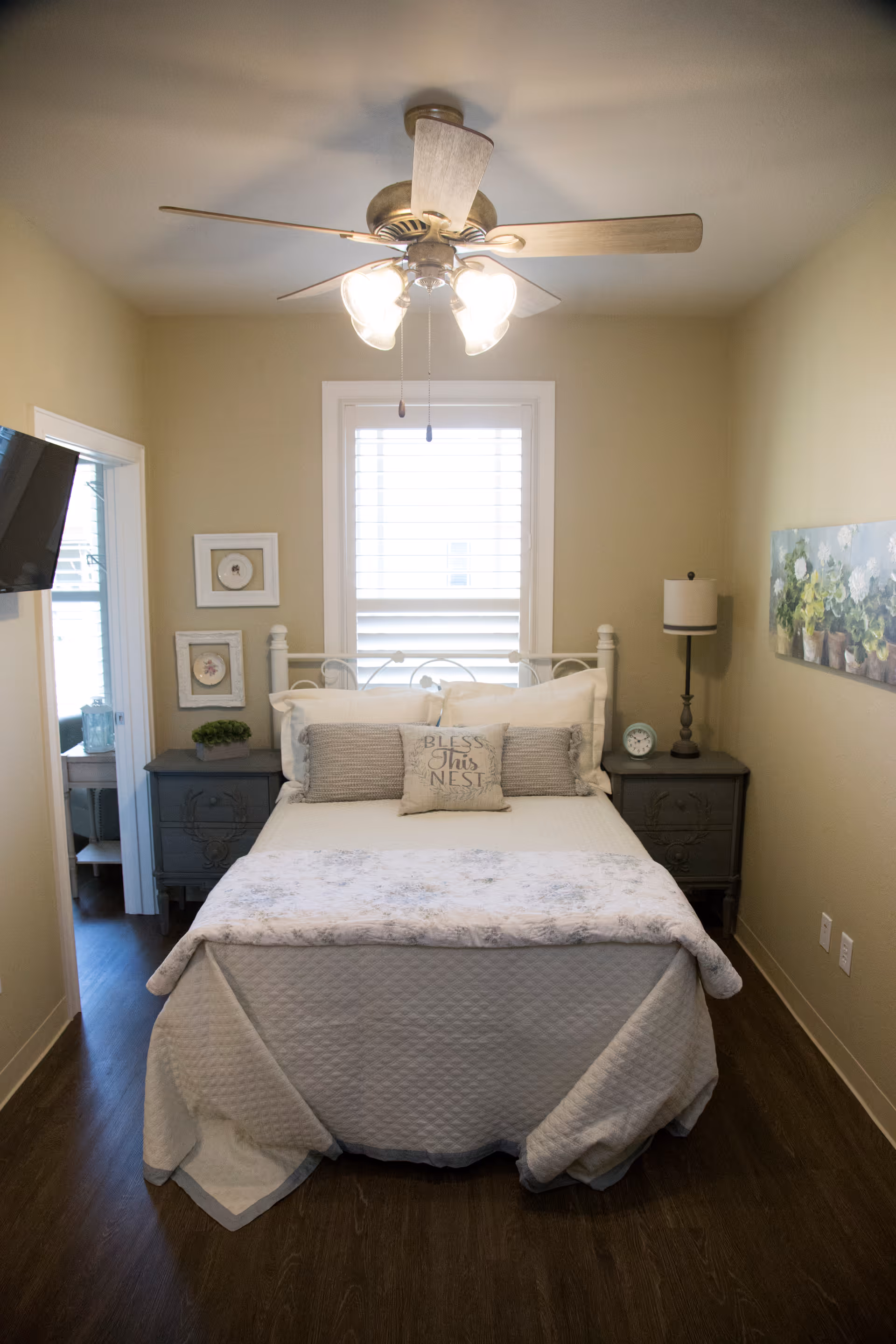 A cozy bedroom with a neatly made bed featuring a decorative pillow that says 'Bless This Nest'. The room has beige walls, a ceiling fan with lights, two matching nightstands with a lamp and clock on the right side, and framed artwork on the walls. A window with white blinds is centered behind the bed, and a flat-screen TV is mounted on the left wall.