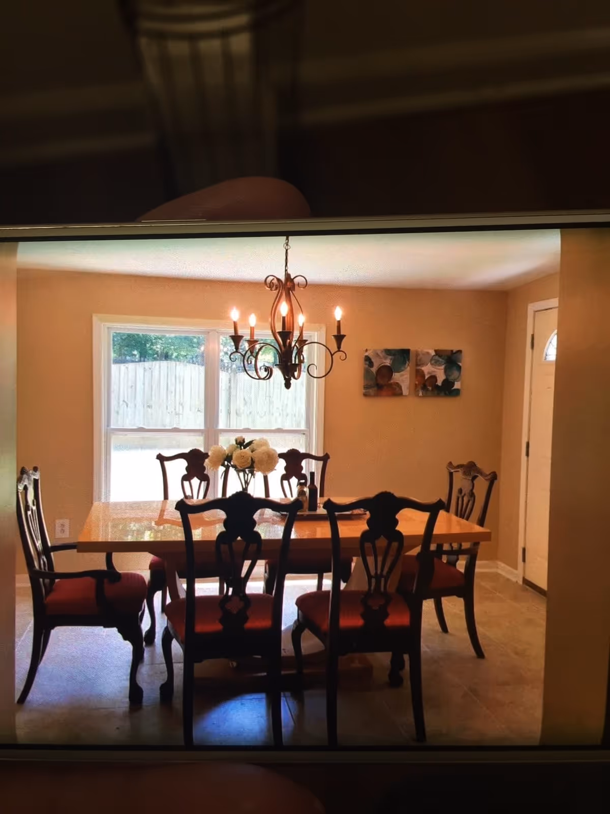 Dining room with a wooden table and six chairs under a decorative chandelier, with a window and doorway in the background.