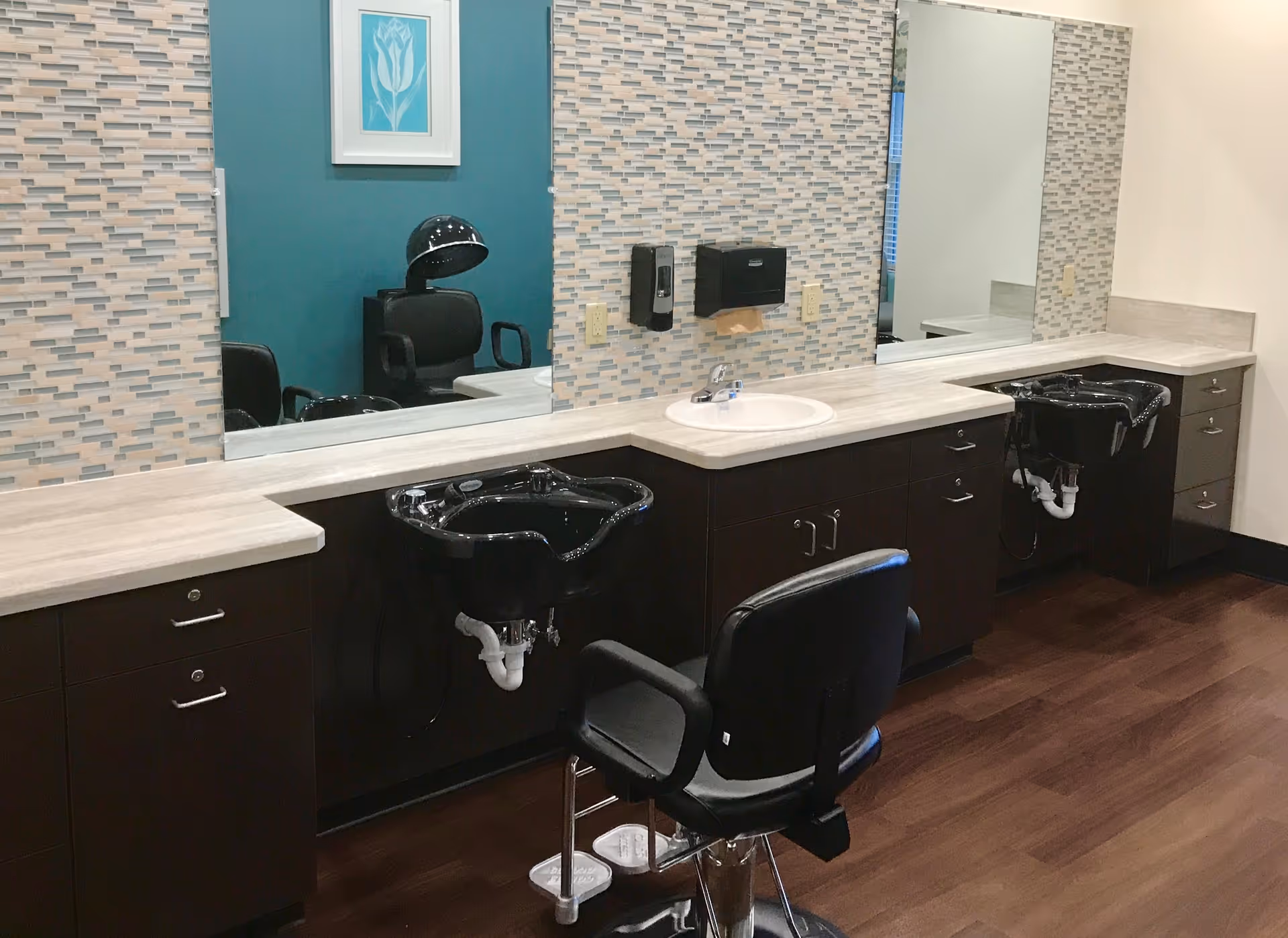 Interior view of a salon area with two black salon chairs in front of black hair washing sinks. The countertop is light-colored with cabinets underneath, and there are large mirrors on the wall with a mosaic tile backsplash. A framed artwork is visible on a teal accent wall in the reflection.