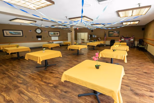 A spacious dining room with multiple square tables covered in yellow tablecloths, each with a small flower pot centerpiece. The room has wood-patterned flooring, beige walls, and ceiling lights with decorative covers. Blue and white streamers hang from the ceiling. There are framed pictures and a clock on the walls, and an American flag is visible in the background.