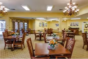 A spacious dining room with multiple wooden tables and chairs arranged neatly. Each table has a floral centerpiece. The room features yellow walls, chandeliers hanging from the ceiling, framed pictures on the walls, and large windows with curtains allowing natural light to enter.
