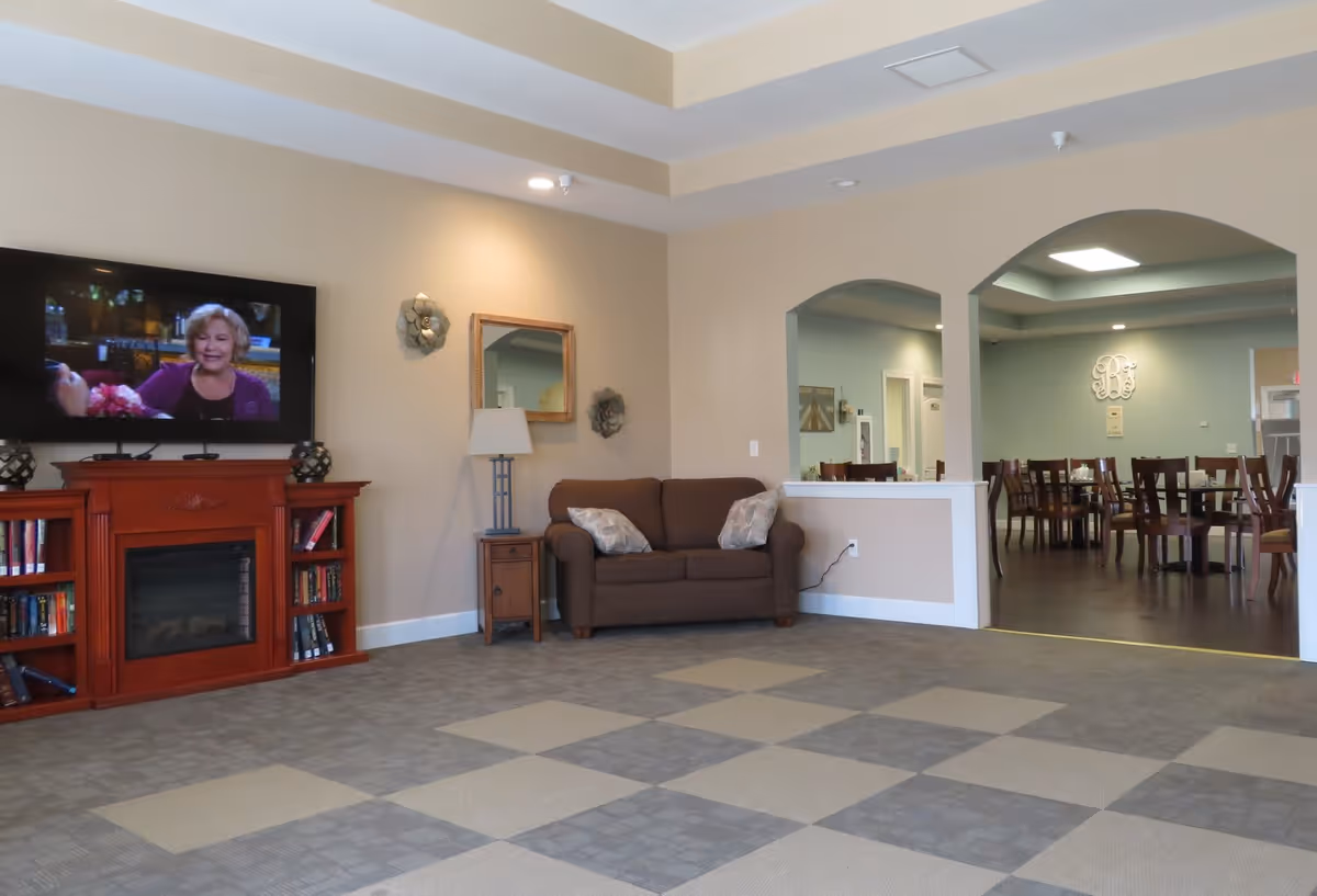 Bright senior living common room with a TV above a fireplace, a sofa, and a view into a dining area through arched openings.