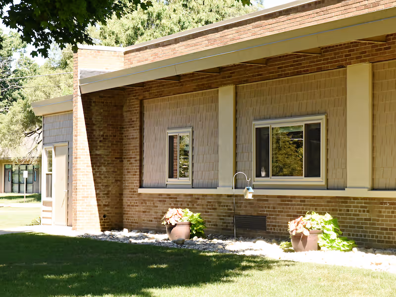 Brick exterior wall of a single-story building with two windows, potted plants, and a grassy lawn.