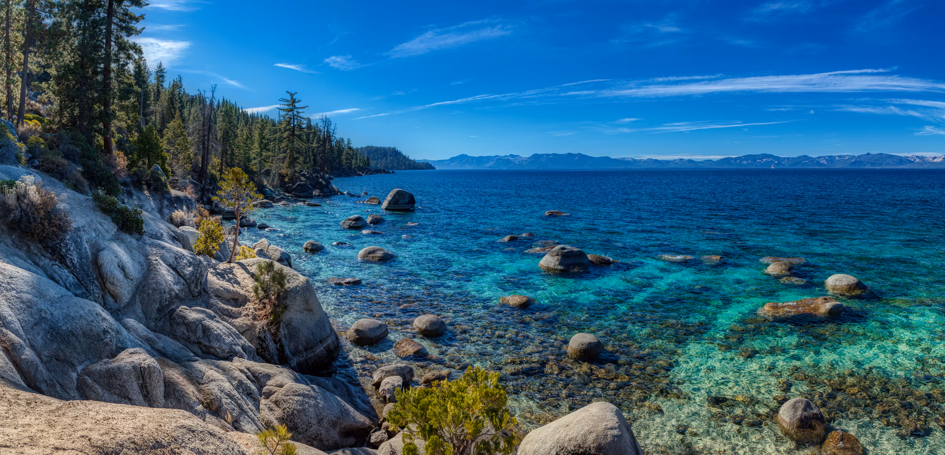 A scenic view of a rocky shoreline with clear turquoise water and large boulders. Pine trees cover the rocky land on the left side, and mountains are visible in the distance under a blue sky with wispy clouds.