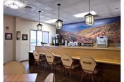 Interior view of a dining area with a long wooden counter and several chairs labeled 'Asbury Place'. The counter has coffee machines and dispensers. Three hanging lantern-style lights illuminate the space, and a large mural of a scenic mountain landscape decorates the wall behind the counter.
