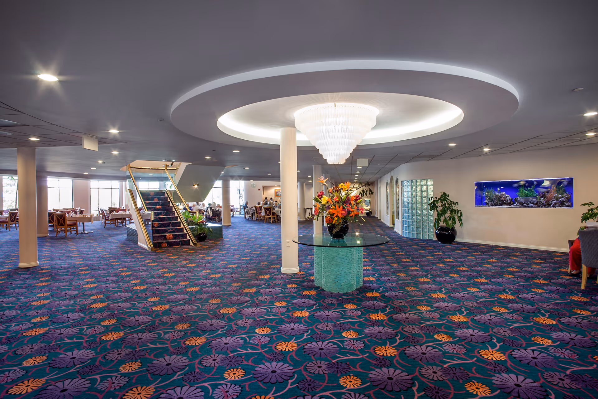 Spacious interior area with a patterned carpet featuring purple and orange flowers, a central round glass table with a floral arrangement, a large chandelier above, a staircase with gold railings, dining tables and chairs in the background, and a wall-mounted aquarium on the right side.