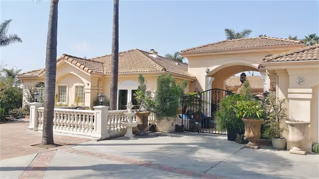 Exterior view of a single-story residential building with a tiled roof, palm trees, and various potted plants near the entrance. The building has a gated archway and a decorative white balustrade along the front.