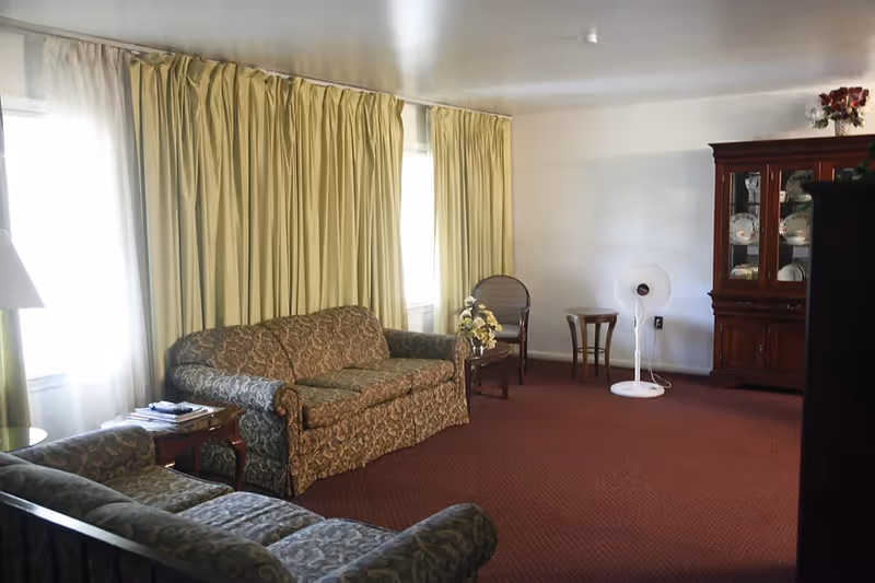 Senior living facility living room with patterned sofas, curtains, a china cabinet, and a fan.
