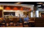 Interior view of a dining area in a senior living facility with patterned upholstered chairs and tables. In the background, there is a food service counter labeled 'The Bistro' with staff behind the counter. The space has warm lighting and a modern design with exposed ceiling elements.