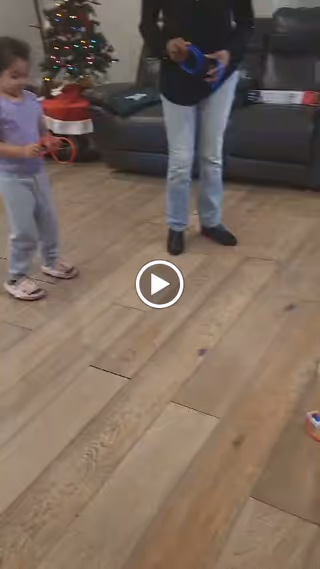 A young girl and an adult standing on a wooden floor in a living room area. The girl is holding a red ring and the adult is holding a blue ring. In the background, there is a decorated Christmas tree with lights and a red tree skirt, and a dark-colored couch with pillows.