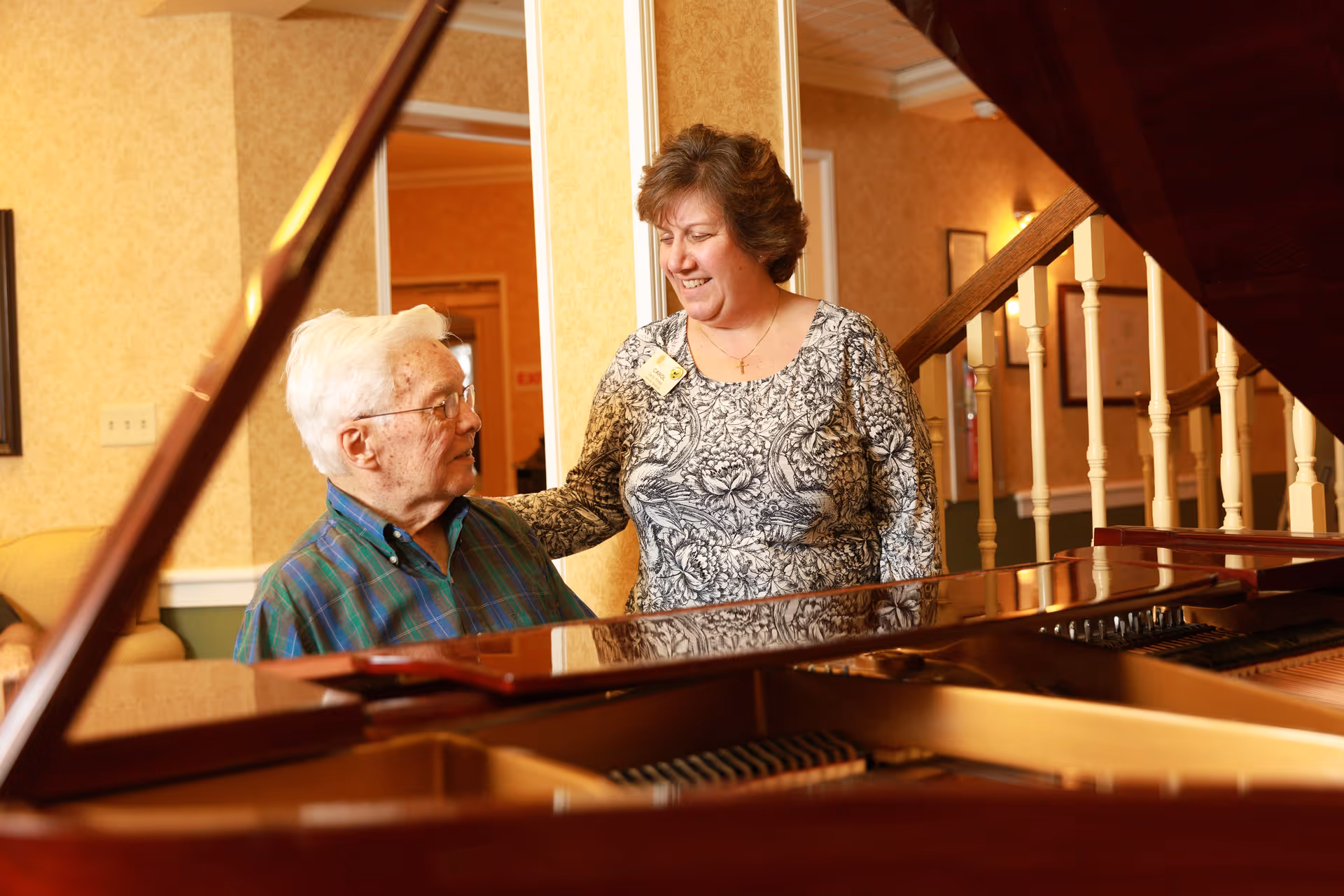 An elderly man sitting at a grand piano is smiling and looking up at a woman standing next to him who is also smiling. They are in a warmly lit room with yellow patterned wallpaper and a staircase with white spindles in the background.