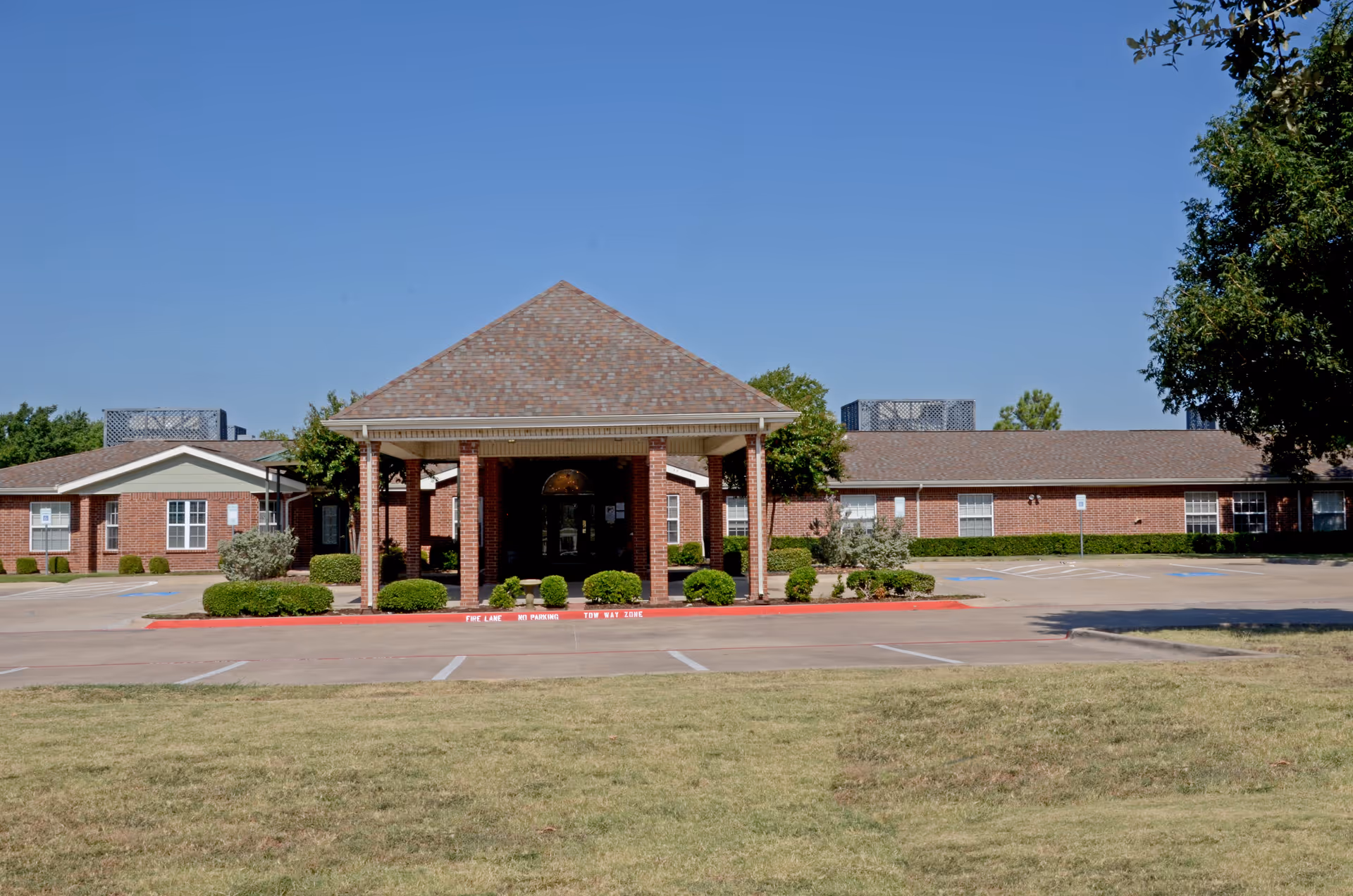 Front exterior of a single-story brick senior living facility with a covered entrance, parking area, and lawn.