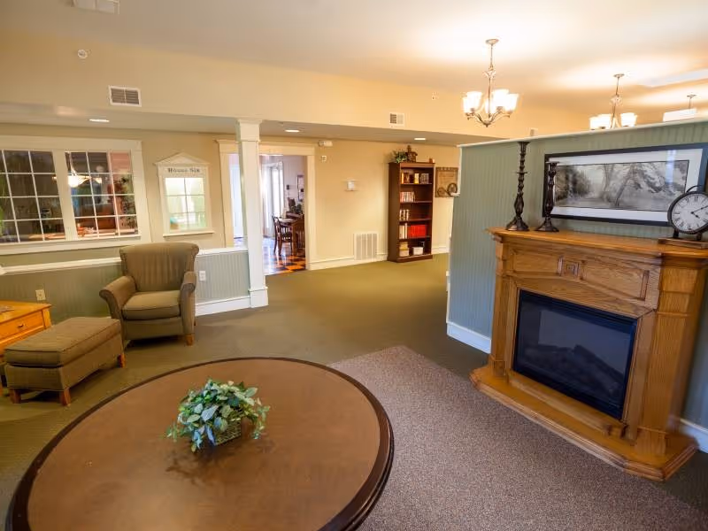 A cozy living room area in Buchanan Meadows facility featuring a round wooden coffee table with a small green plant centerpiece, a comfortable armchair with matching ottoman, a wooden electric fireplace with decorative candlesticks and a framed picture above it, and a bookshelf in the background. The room has beige walls, carpeted floor, and warm lighting from ceiling fixtures.