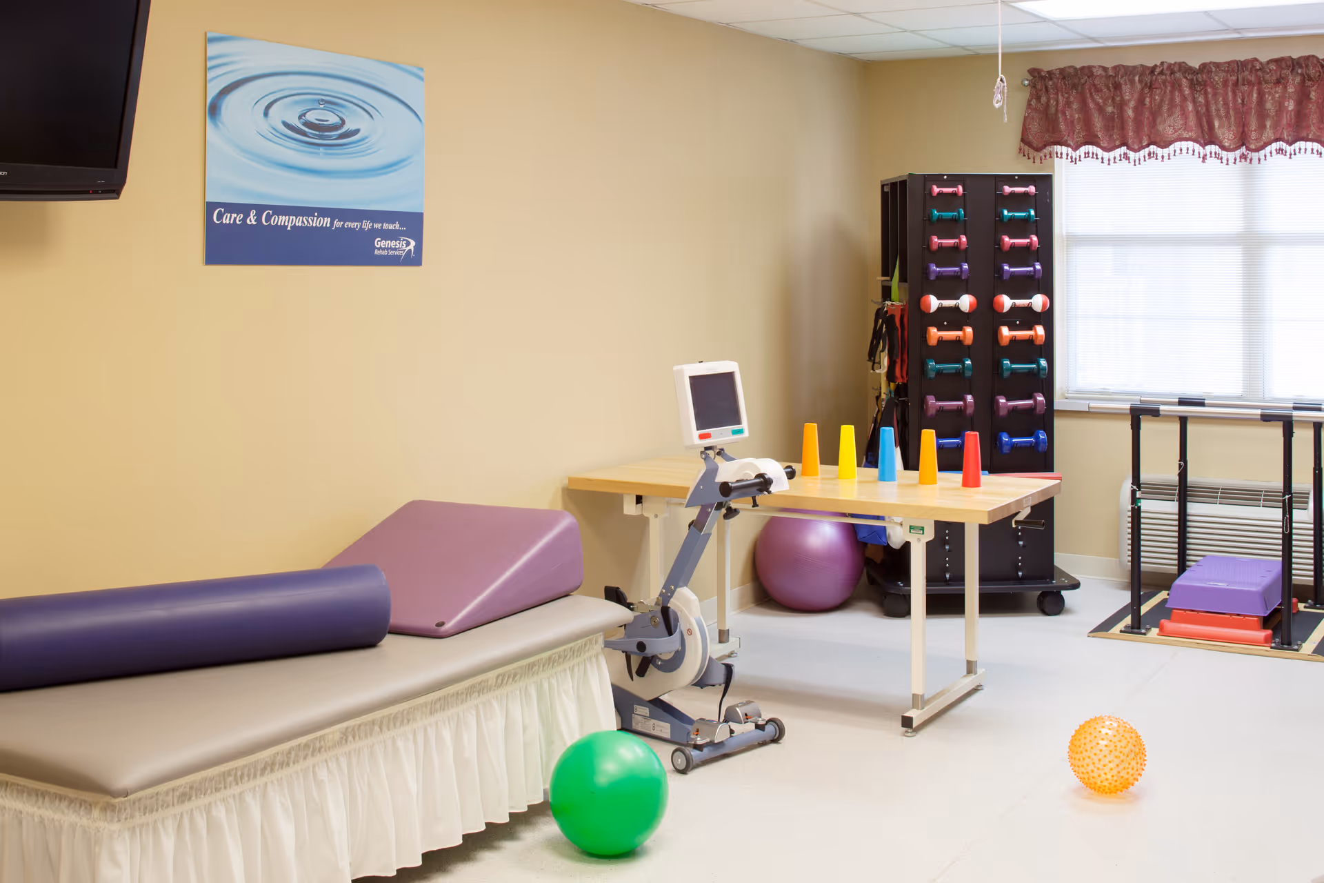 A physical therapy or rehabilitation room with exercise equipment including a stationary bike, colorful cones on a table, various dumbbells on a rack, exercise balls, and padded therapy tables. A poster on the wall reads 'Care & Compassion for every life we touch... Genesis'.