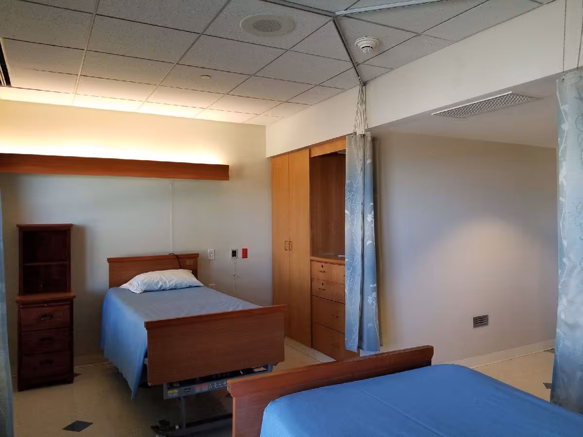 A simple, clean room with two single beds covered in blue bedspreads. There is a wooden nightstand with drawers and a small shelving unit next to one bed. The room has a ceiling with recessed lighting and a curtain divider between the beds. A wooden built-in cabinet with drawers is visible in the background.