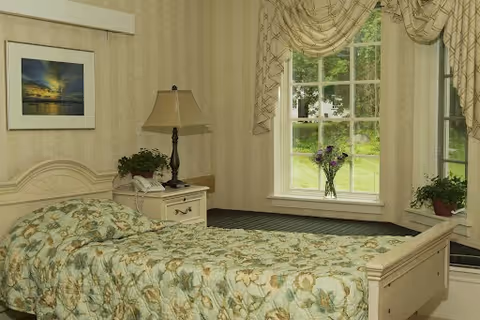 A cozy bedroom with a single bed covered in a floral patterned bedspread. Next to the bed is a nightstand with a lamp, a telephone, and a small potted plant. A framed picture hangs on the wall above the bed. A window with decorative curtains lets in natural light and shows greenery outside. Another potted plant is on the window sill.