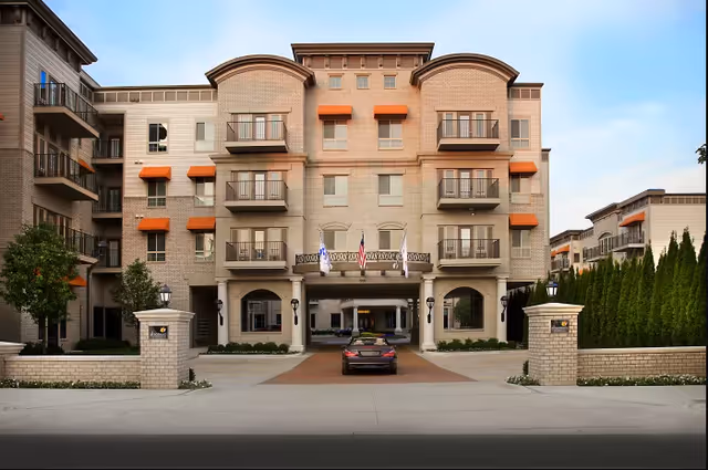 Front exterior view of a multi-story senior living facility building with beige brick walls, multiple balconies with black railings, and orange awnings above some windows. There is a driveway leading to the entrance with a car parked under a covered area. Two flagpoles with flags are positioned above the entrance. The building is surrounded by trees and shrubs.