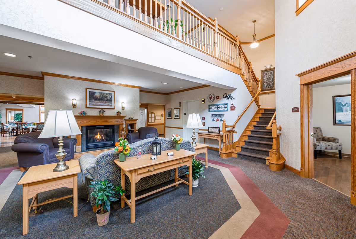 A cozy senior living facility common area with a fireplace, armchairs, a sofa, wooden tables with lamps and flower arrangements, a staircase with wooden railing leading to an upper floor, and wall decorations including a clock and coffee-themed signs.
