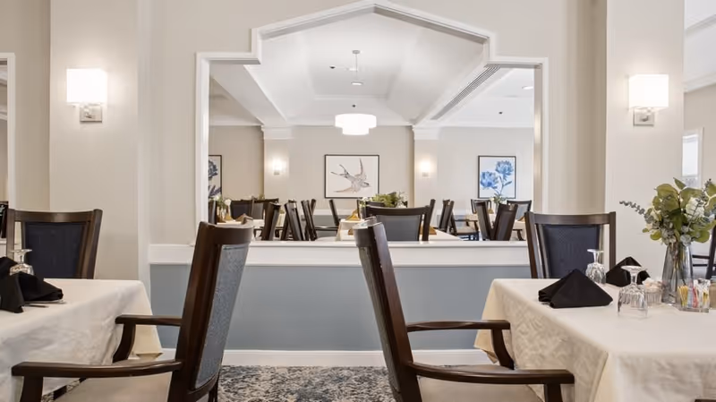 A bright and elegant dining room in Aspenwood Senior Living Community featuring tables covered with white tablecloths, dark wooden chairs with armrests, black folded napkins, glassware, and floral centerpieces. The room has soft beige walls, wall sconces, and framed artwork of birds and flowers. A large mirror reflects the room, enhancing the spacious feel.
