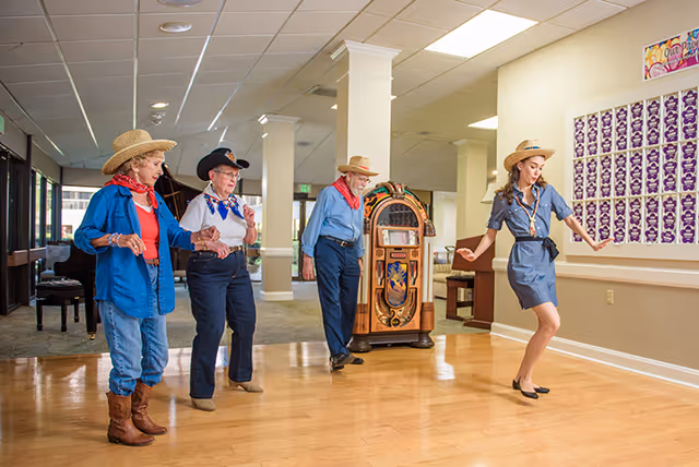 Four people dressed in cowboy hats and western attire are dancing on a wooden floor inside a well-lit room with columns, a jukebox, and a piano in the background.