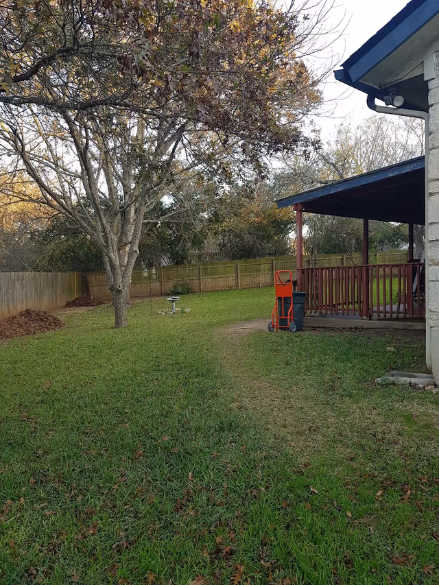 Grassy fenced backyard with a large tree, a birdbath, and a covered porch with a hand truck near the house.