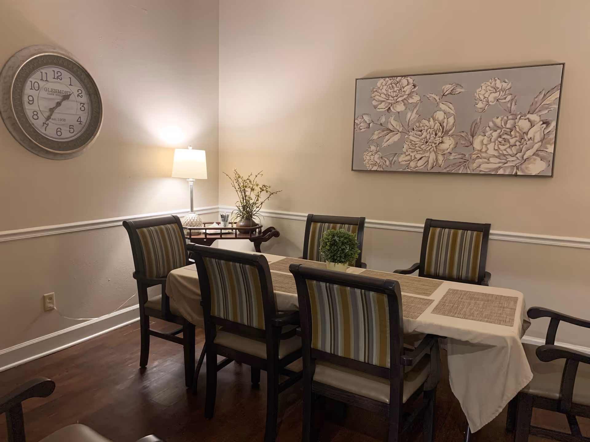 Dining room with a rectangular table covered by a tablecloth and placemats, six striped chairs, a wall clock, lamp, and floral wall art.