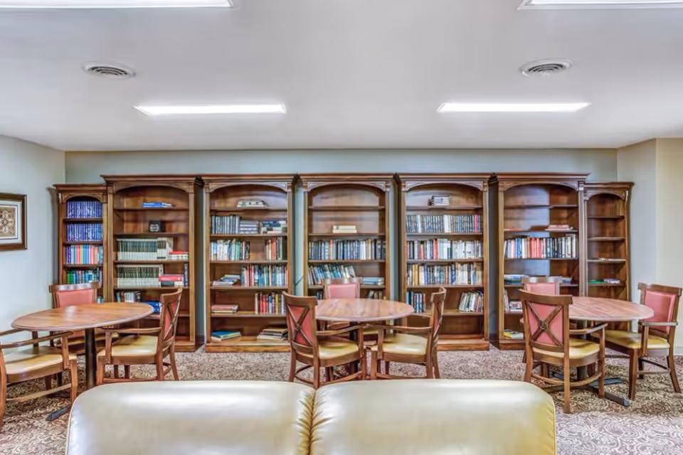 A cozy library room in a retirement community featuring five wooden bookshelves filled with books against a wall. In front of the bookshelves are three round wooden tables, each surrounded by four wooden chairs with cushioned seats and backs. The room has a patterned carpet and a white ceiling with recessed lighting.