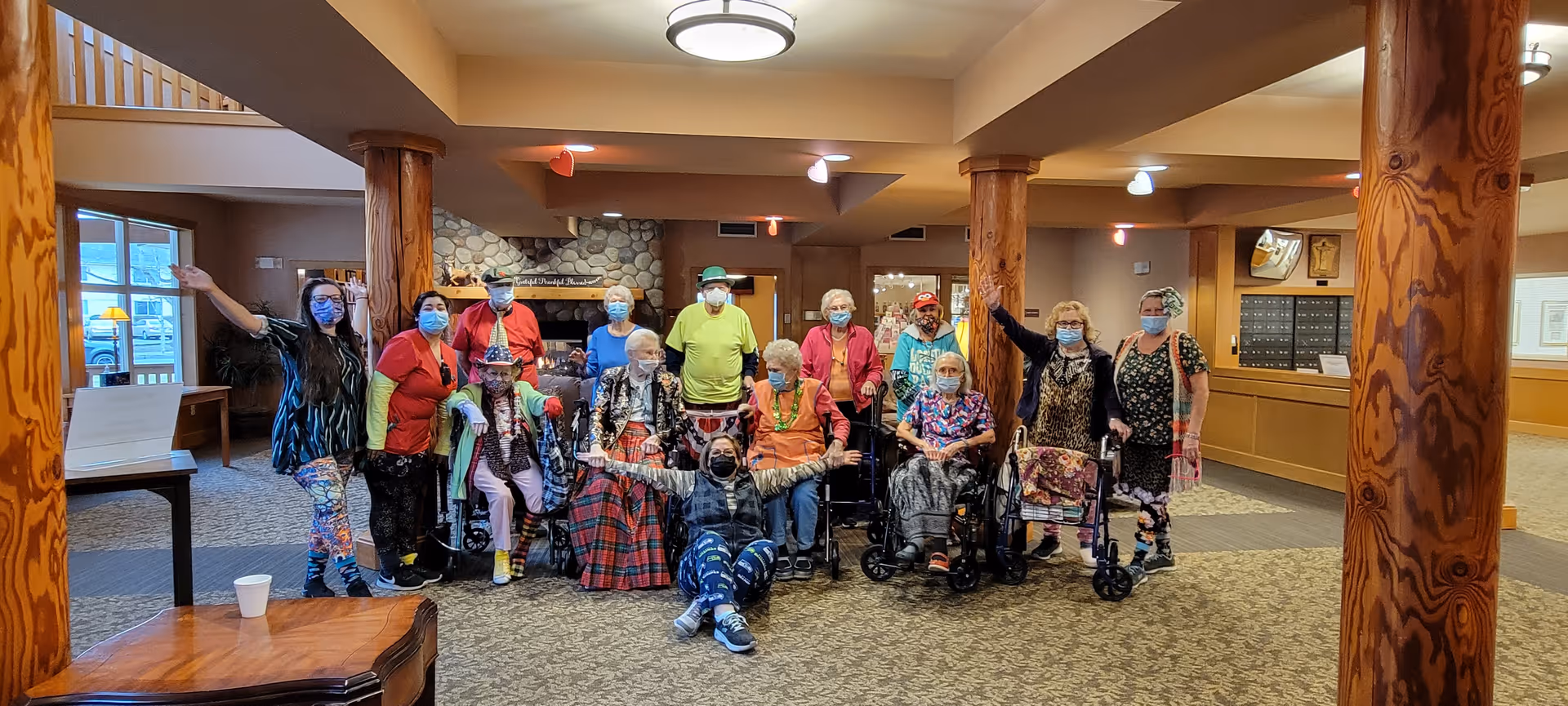 A group of masked people posing together in a community room with wooden columns and a stone fireplace.
