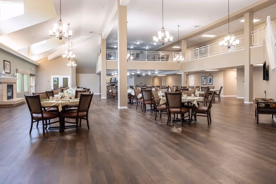 Spacious dining area with multiple tables and chairs arranged neatly, set with tablecloths, cups, and plates. The room features high ceilings with several chandeliers, large windows allowing natural light, a fireplace on the left, and a second-floor balcony overlooking the space.