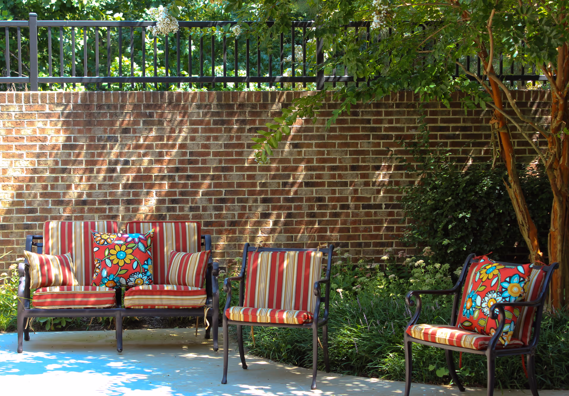 Outdoor patio with striped cushioned chairs and colorful floral pillows set against a brick wall and garden greenery.