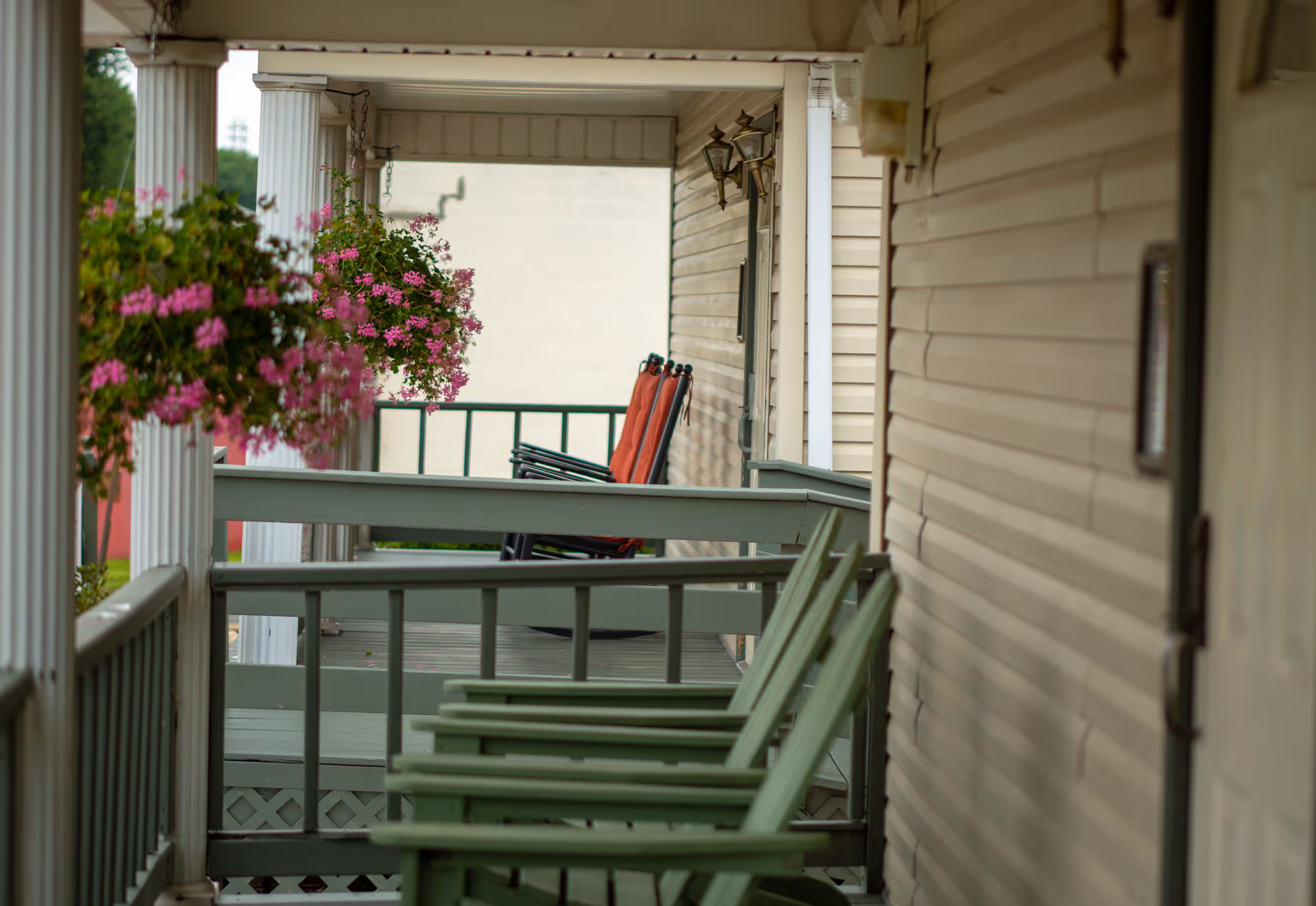 A covered porch area with green wooden rocking chairs and hanging baskets of pink flowers. The porch has white columns and beige siding on the building wall.