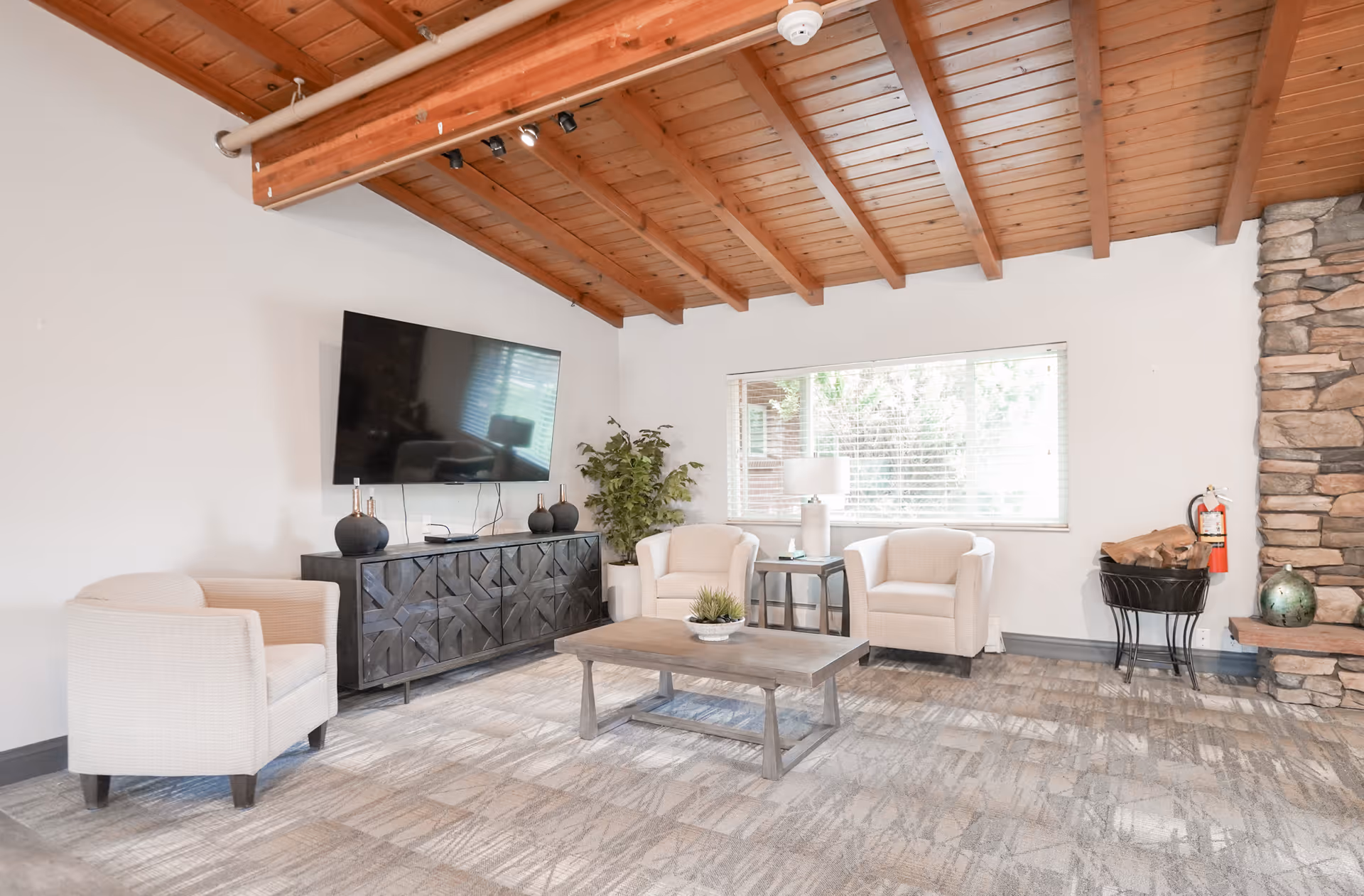A cozy living room with a wooden vaulted ceiling, a large flat-screen TV mounted on the wall above a dark wooden cabinet, three beige armchairs arranged around a wooden coffee table, a large window with blinds, a stone fireplace with a firewood holder and fire extinguisher nearby, and a potted plant in the corner.