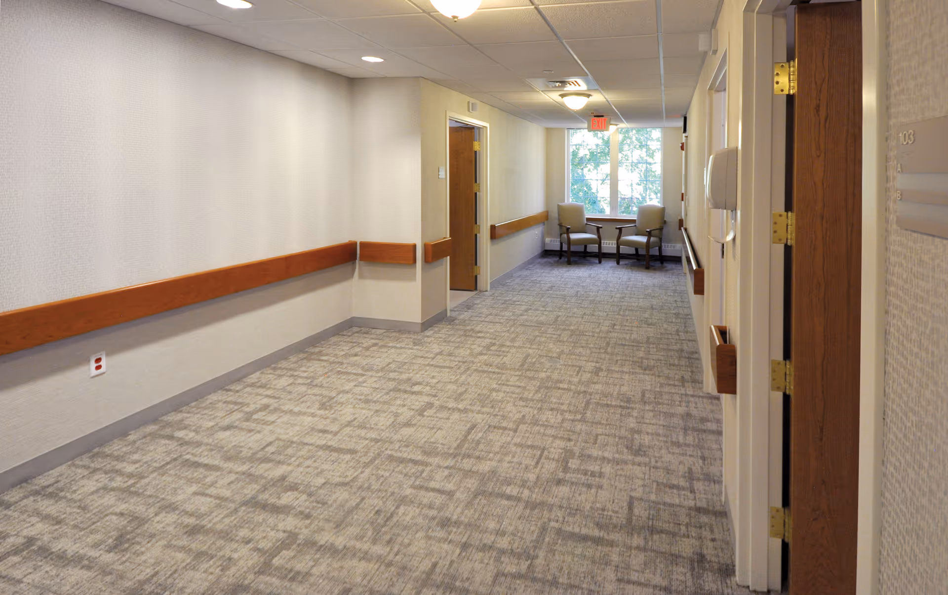 A clean, well-lit hallway in a healthcare facility with beige walls, carpeted floor, wooden handrails on both sides, and two chairs with a small table near a window at the far end.