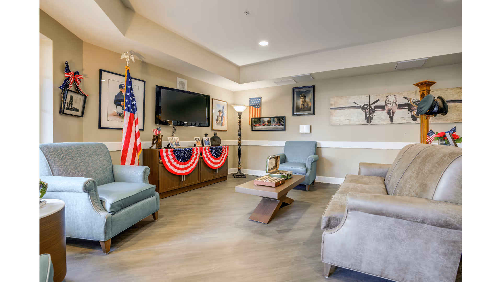 A cozy living room with light blue and beige armchairs and a beige sofa arranged around a modern wooden coffee table. The room is decorated with patriotic American flags and red, white, and blue bunting. On the walls are framed pictures, including portraits and an airplane artwork. A flat-screen TV is mounted above a wooden cabinet, and a tall floor lamp stands in the corner.