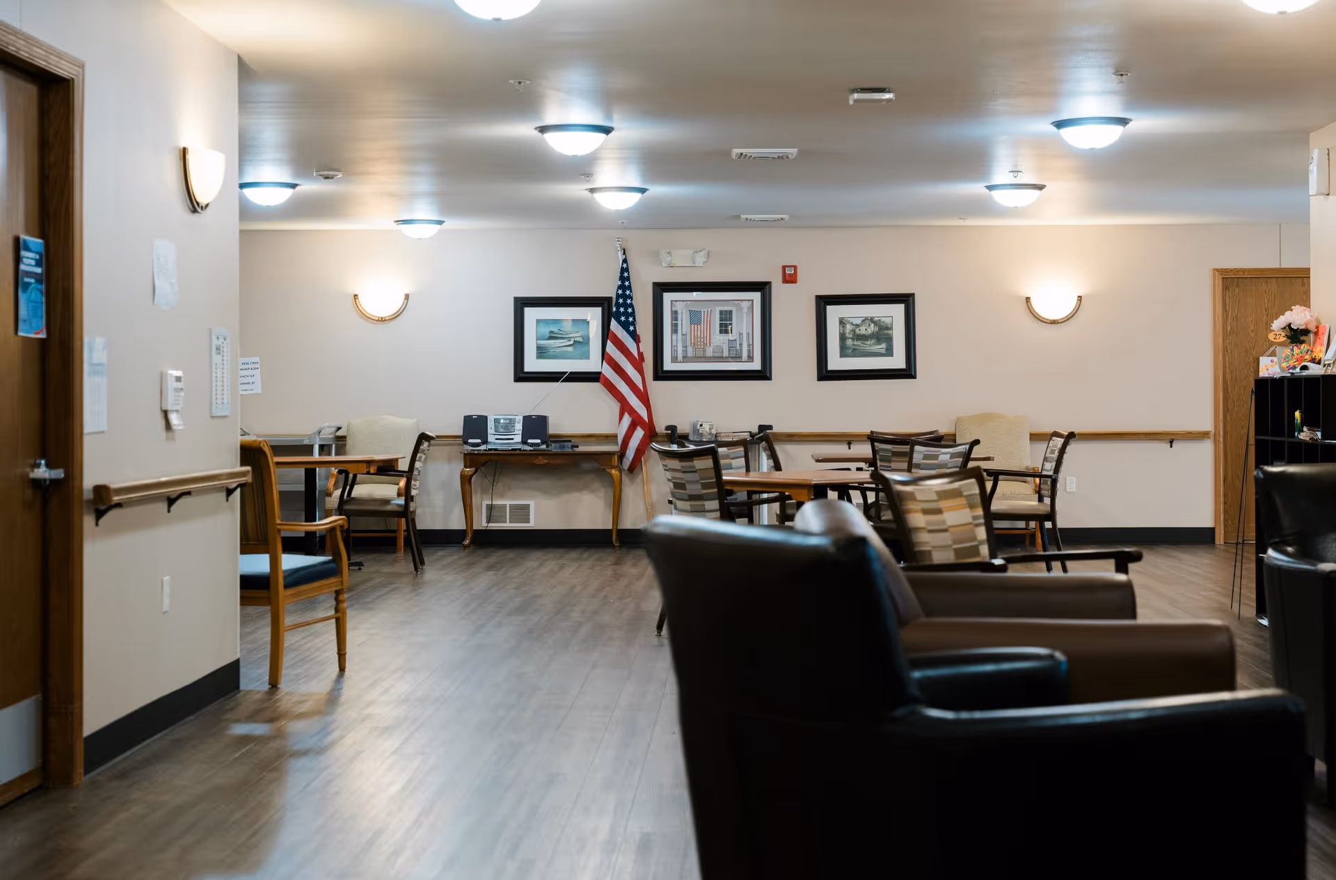 Interior view of a common area in an assisted living facility with wooden flooring, several tables and chairs arranged for seating, an American flag, framed pictures on the wall, and soft lighting from ceiling fixtures and wall sconces.