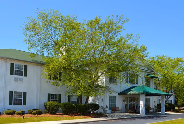 Two-story white senior living building with a green roof and covered front entrance surrounded by trees and shrubs.