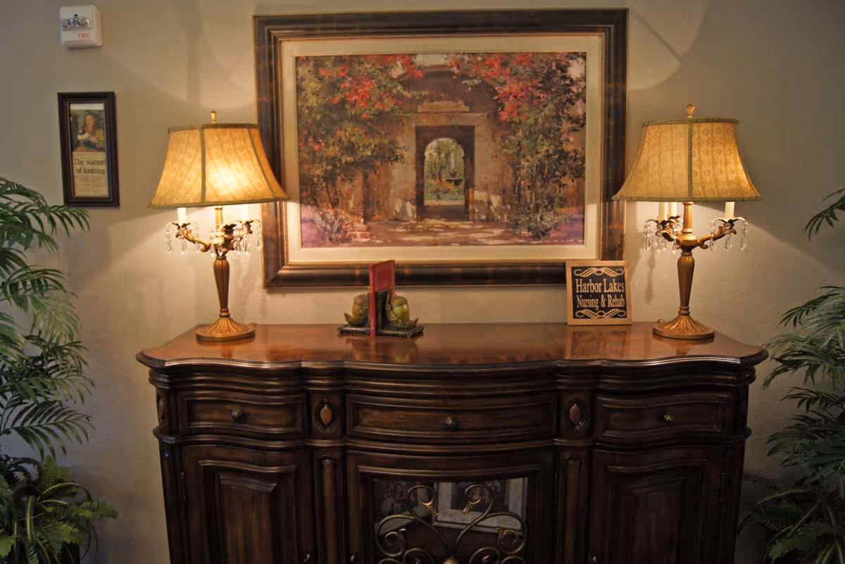 A wooden sideboard with two ornate table lamps on either end, a framed painting of an archway with foliage above it hanging on the wall behind, and a small sign that reads 'Harbor Lakes Nursing & Rehab' placed on the right side of the sideboard. There are also some decorative items and plants on either side of the sideboard.