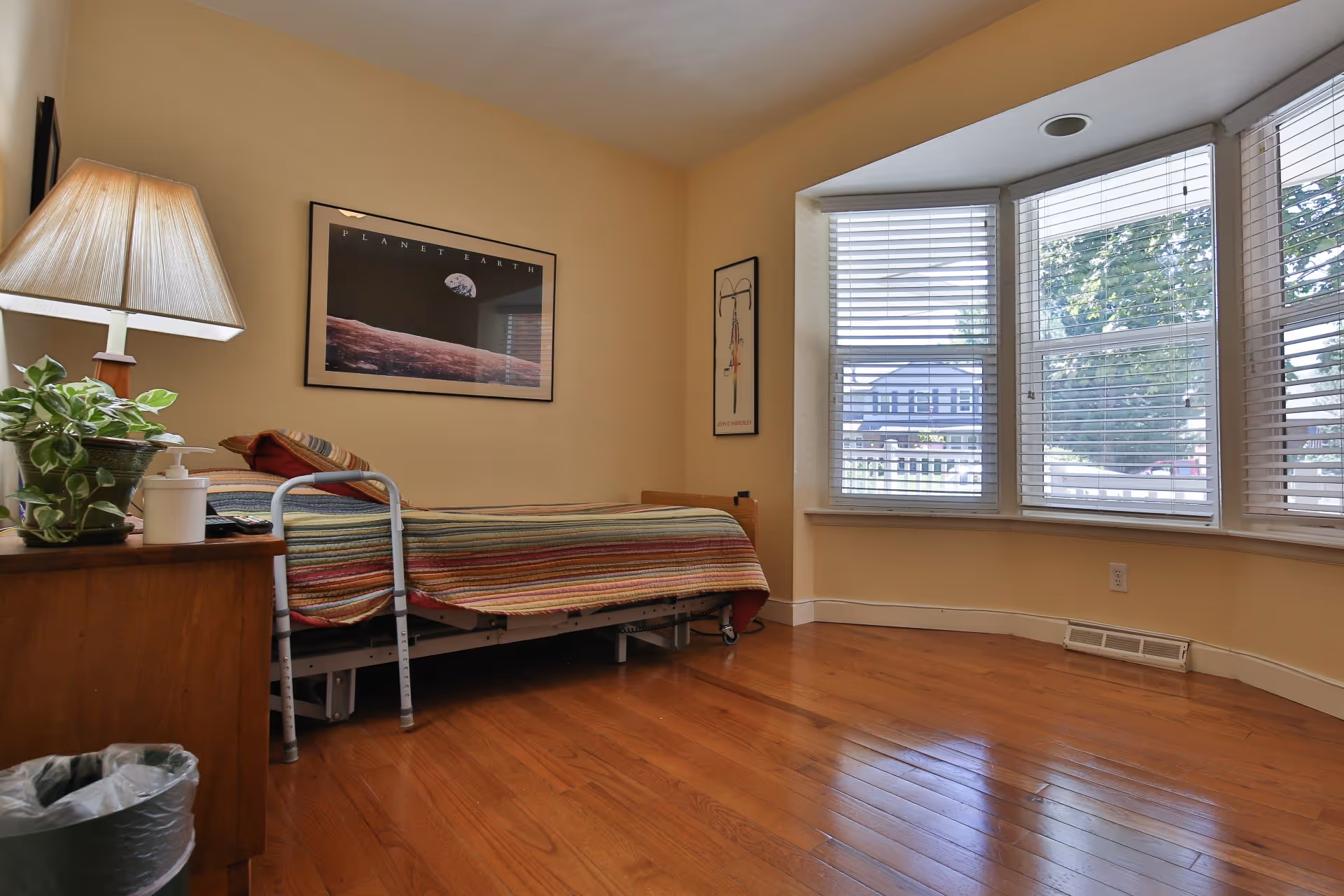 A small bedroom with a single bed covered with a striped blanket. There is a wooden nightstand with a lamp, a plant, and a soap dispenser. A walker is positioned next to the bed. The room has wooden floors and a large bay window with white blinds, letting in natural light. Two framed pictures hang on the beige walls.
