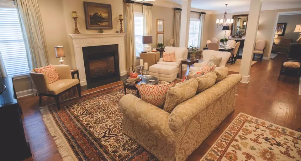 A cozy living room with a patterned beige sofa, two white armchairs, a wooden coffee table with candles and decor, a fireplace with a painting above it, and a dining area with a table and chairs in the background. The room has wooden floors, area rugs, and windows with curtains allowing natural light.