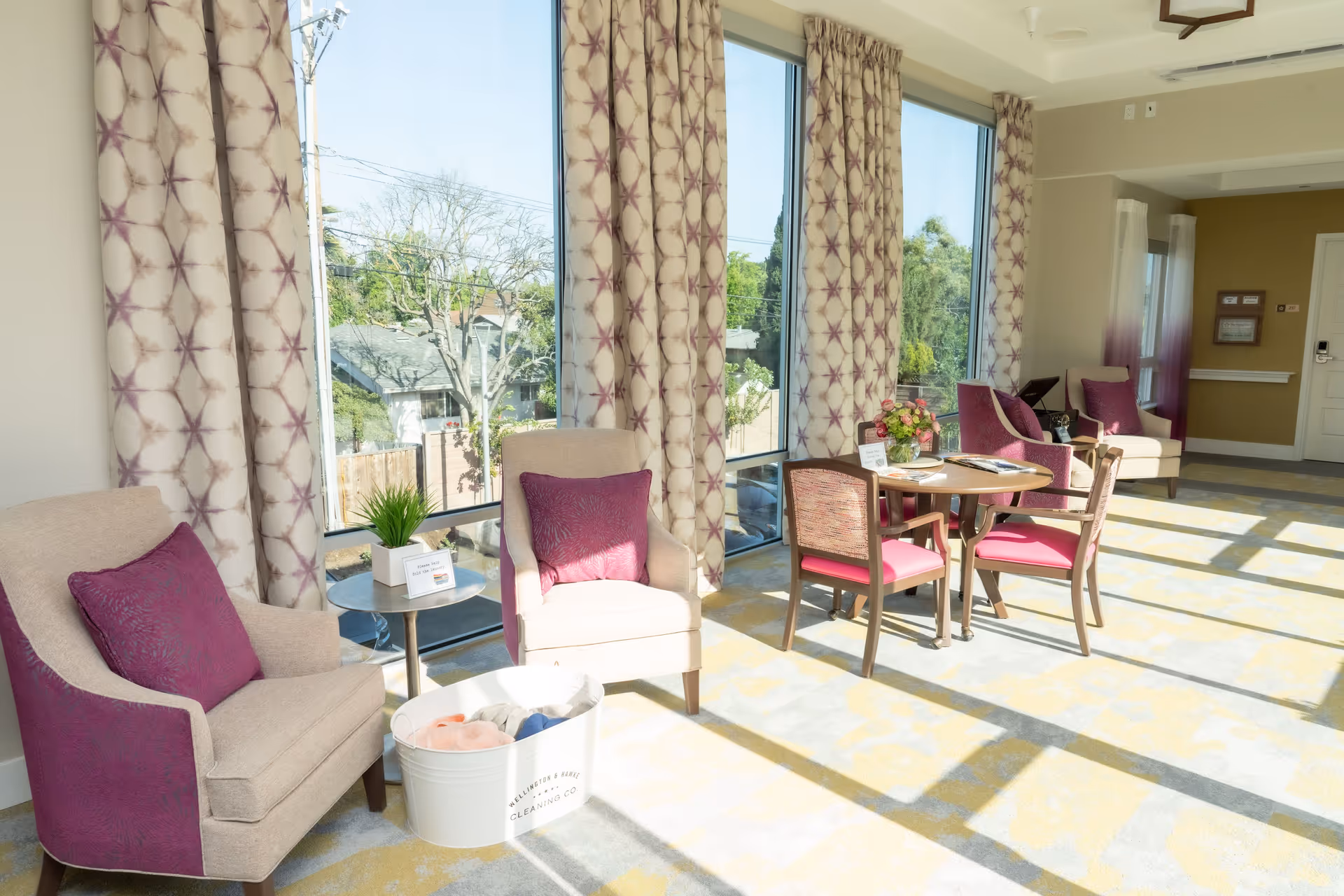 A bright and airy sitting area with large windows letting in natural light. The room features beige armchairs with purple cushions, a small round table with two chairs, and patterned curtains. Outside the windows, trees and neighboring houses are visible. The carpet has a yellow and gray geometric pattern, and there is a white bucket labeled 'Wellington & Harte Cleaning Co.' on the floor.