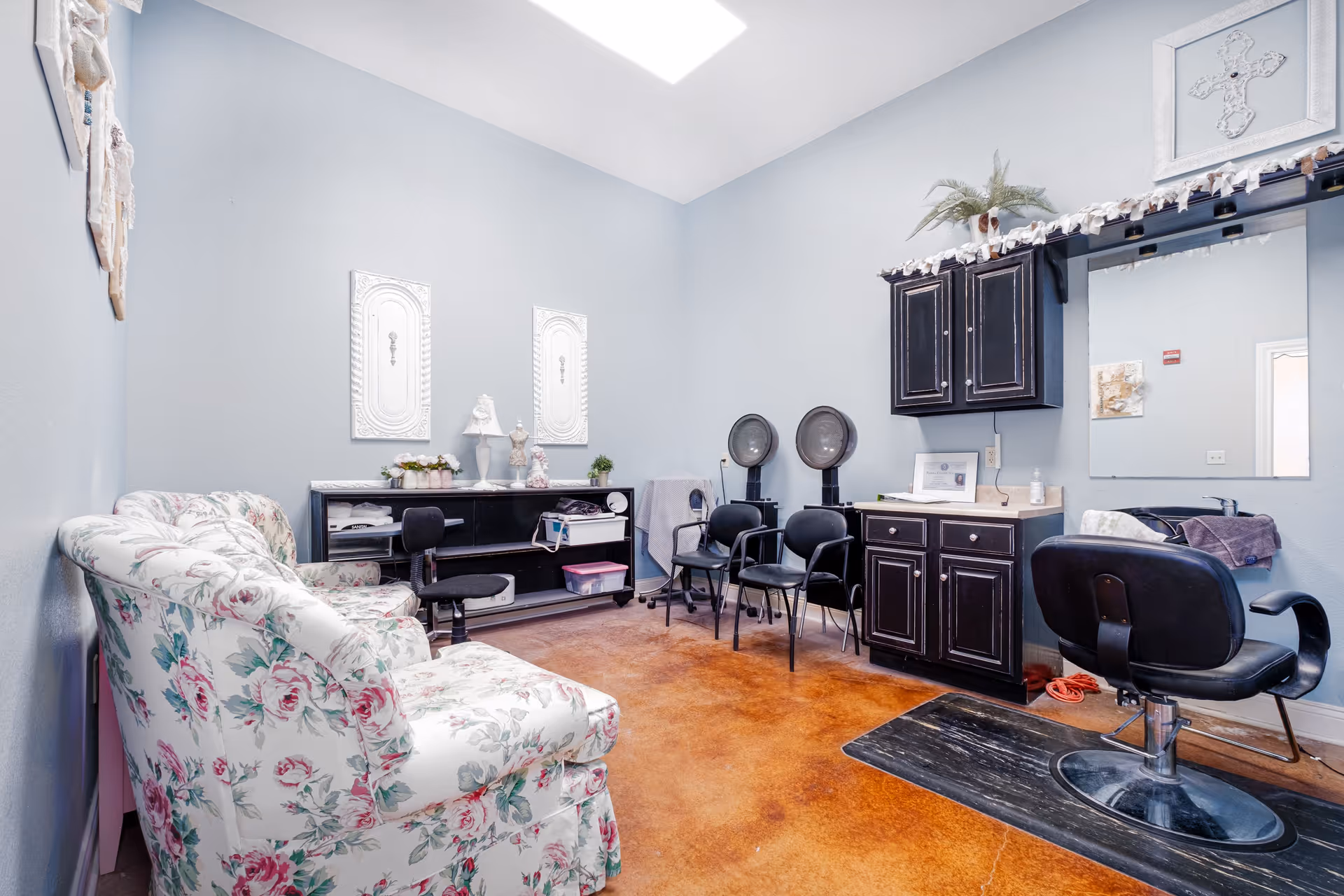 A bright room with light blue walls and a skylight, featuring a floral patterned couch on the left, a black shelving unit with decorative items, three black chairs, two hair drying stations, a black salon chair in front of a large mirror, and black cabinets with a countertop. The floor is a polished brown concrete.