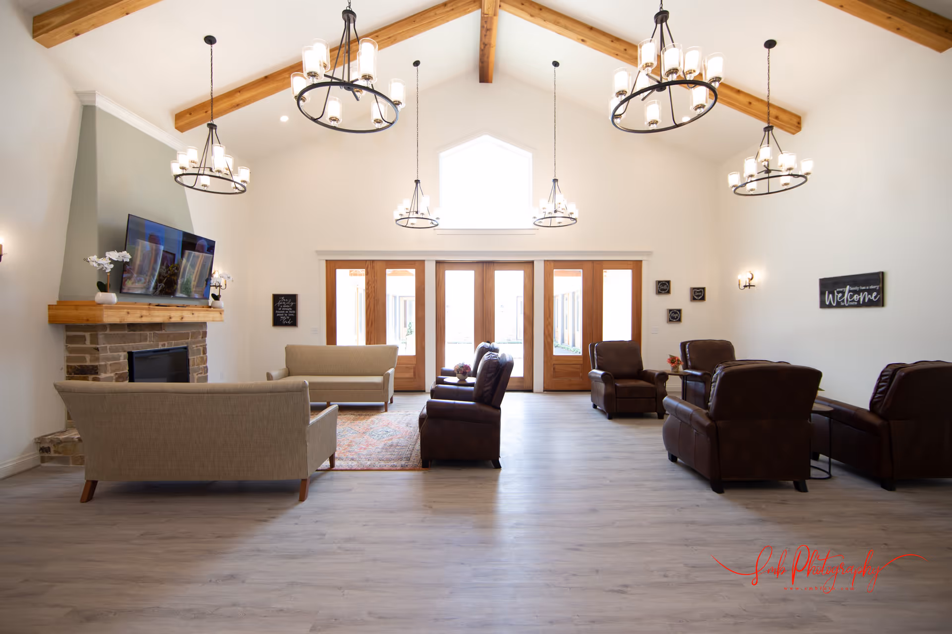 A spacious and bright living room with high vaulted ceilings featuring exposed wooden beams and multiple modern chandeliers. The room has light-colored walls and flooring, with a stone fireplace on the left side topped with a wooden mantle and a mounted flat-screen TV. There are two beige sofas facing the fireplace and several dark brown leather armchairs arranged around small tables. Large wooden-framed glass doors and a window above them allow natural light to fill the room. Wall decorations include small framed signs and a welcome sign.