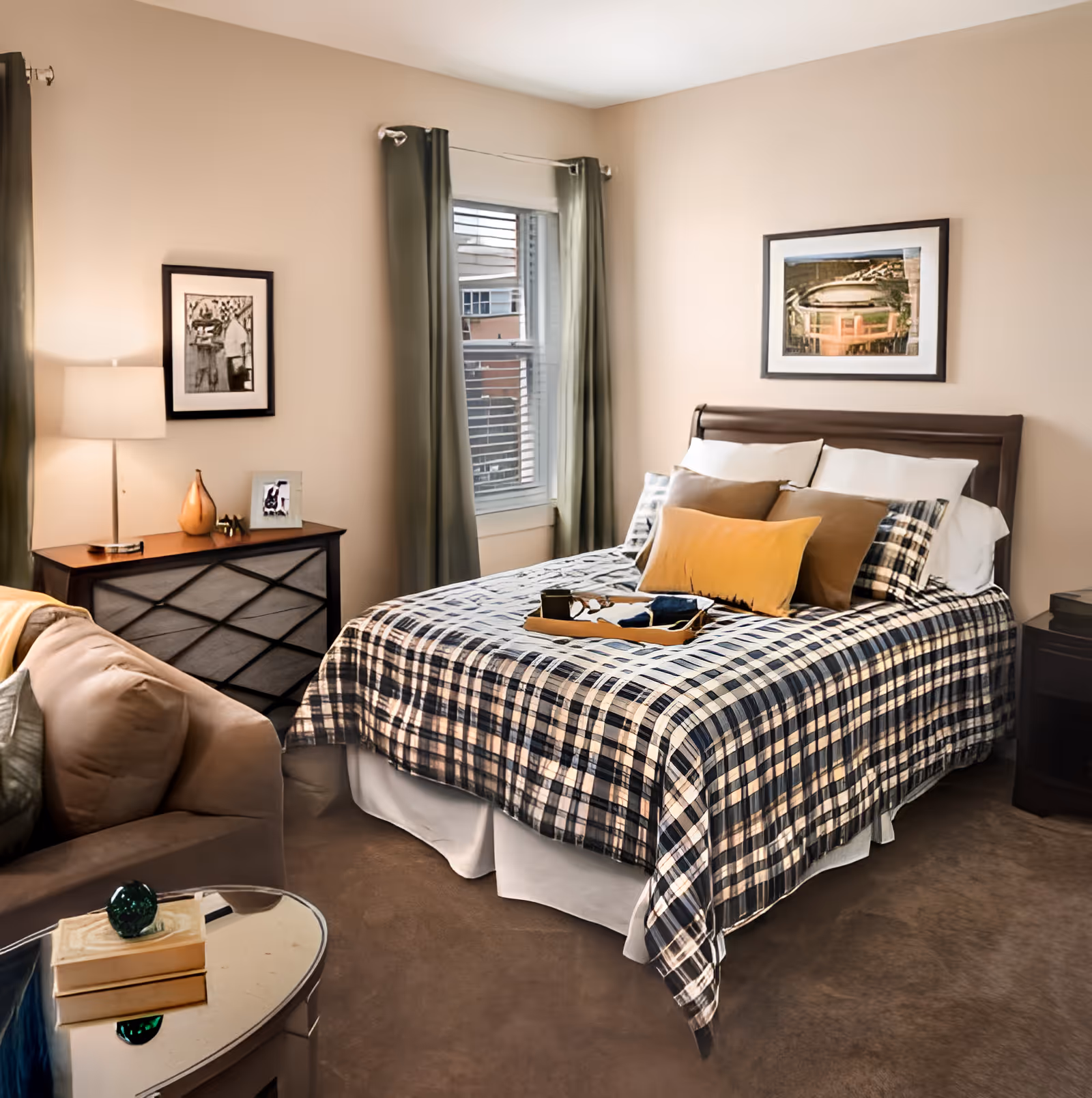 A cozy bedroom featuring a bed with a plaid bedspread and multiple pillows, a wooden nightstand with a lamp and decorative items, a window with green curtains, a framed picture on the wall above the bed, and a beige armchair next to a small round table with books and a decorative object.