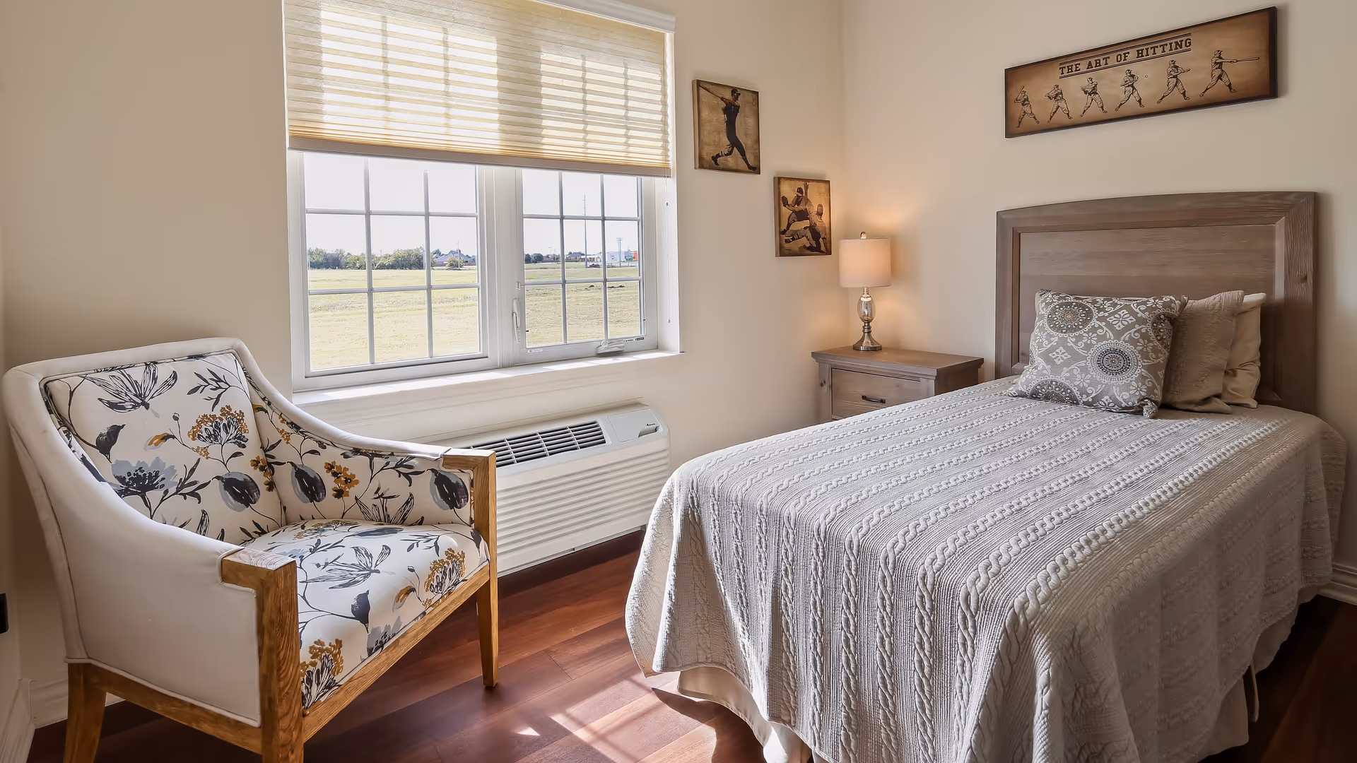 A cozy bedroom with a single bed covered in a textured white blanket and two decorative pillows. Next to the bed is a wooden nightstand with a lamp. A floral upholstered armchair is positioned near a large window with blinds, letting in natural light. The walls are decorated with three framed pictures related to baseball.