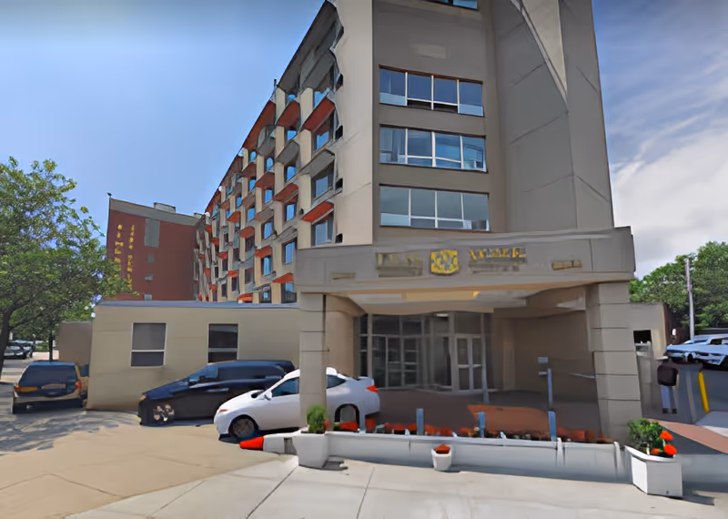 Exterior view of a multi-story assisted living facility building with a covered entrance, several parked cars, and some greenery around the parking area under a clear blue sky.