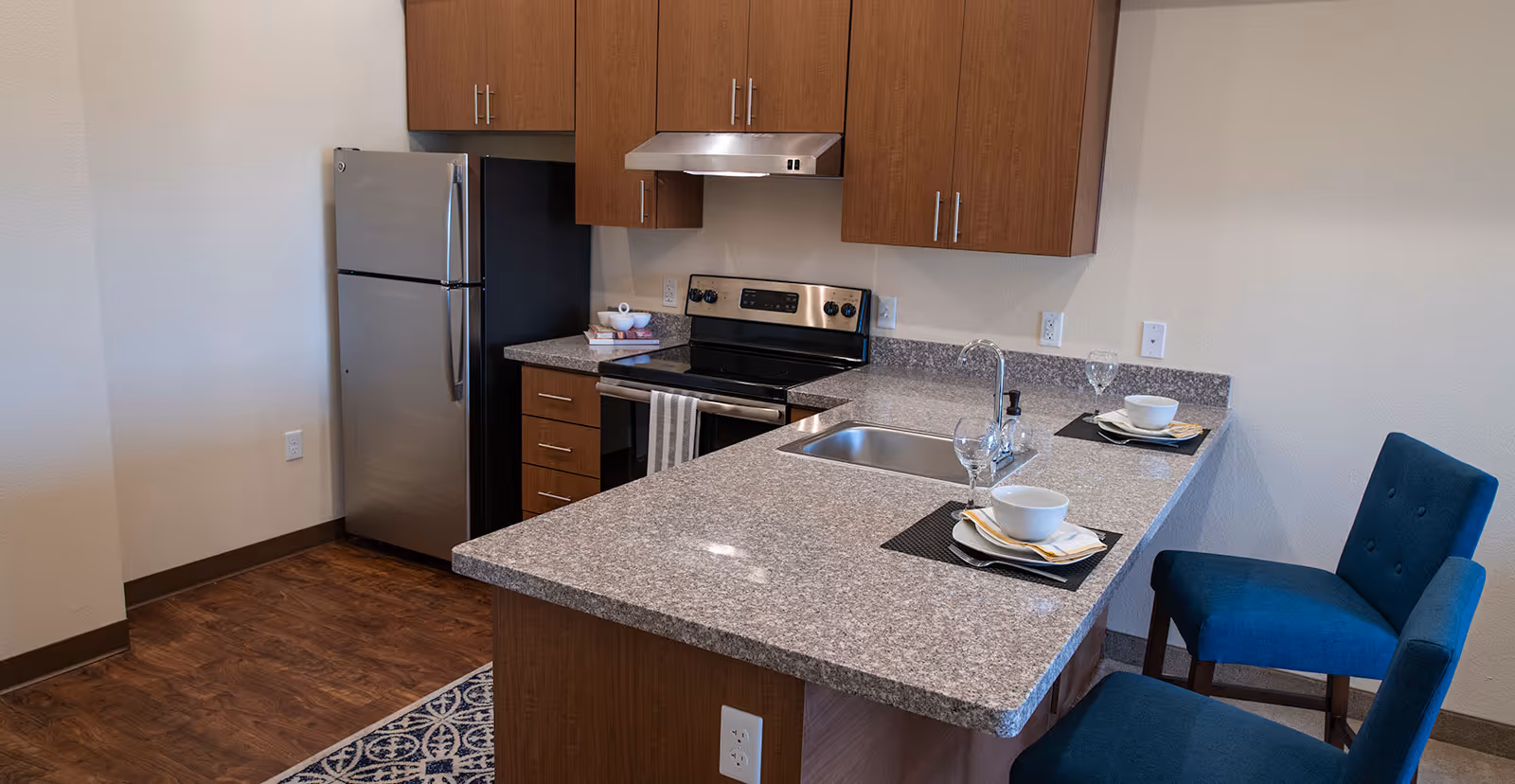Modern kitchen area with granite countertops, stainless steel refrigerator and stove, wooden cabinets, a sink with a faucet, and two blue upholstered chairs at the counter set with bowls, plates, napkins, and wine glasses.